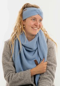 Donna con lunghi capelli mossi, che indossa un cerchietto azzurro di lana lavorata a maglia e una sciarpa abbinata, sorridendo e vestita con un cappotto grigio a trama.