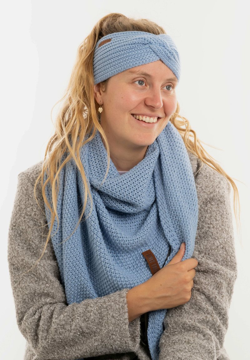 Donna con lunghi capelli mossi, che indossa un cerchietto azzurro di lana lavorata a maglia e una sciarpa abbinata, sorridendo e vestita con un cappotto grigio a trama.