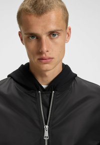 Veste aviateur noire avec une capuche, dotée d'une fermeture éclair argentée, de poignets côtelés et d'une finition lisse et brillante. Le visage du mannequin est visible.