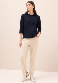 Felpa con cappuccio blu navy con finitura testurizzata, abbinata a pantaloni beige chiaro e scarpe da ginnastica bianche con dettagli bordeaux.