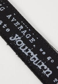 Zwarte stoffen band met witte tekst. Het ontwerp bevat de woorden "JOUW BEURT" en "gemiddeld," met een verouderde textuur en onregelmatige letters.