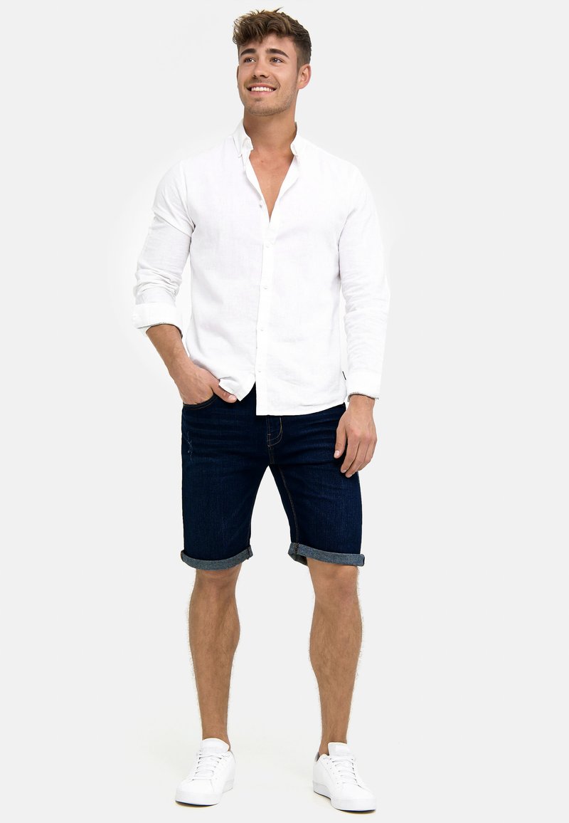 Junger Mann lächelt, trägt ein weißes Langarmhemd mit aufgekrempelten Ärmeln, dunkle Jeansshorts und weiße Turnschuhe, steht mit einer Hand in der Tasche.