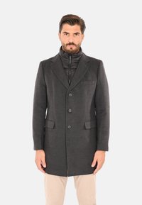 Uomo con cappotto grigio scuro abbottonato sopra una giacca imbottita nera e pantaloni beige, in piedi davanti a uno sfondo chiaro e uniforme.