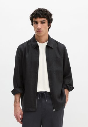 Junger Mann mit lockigem Haar, der ein schwarzes Zip-Up-Hemd über einem weißen T-Shirt und schwarze Hose mit Kordelzug trägt, stehend vor einem schlichten weißen Hintergrund.