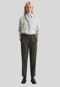 Femme blonde se tenant avec les mains dans les poches de son pantalon gris foncé, portant une chemise à boutons bleu clair sur un col roulé assorti et des chaussures noires.