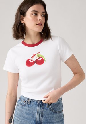 T-shirt blanc avec col en rib rouge, orné d'un graphique de deux cerises avec une feuille et une petite étiquette, associé à un jean en denim bleu.
