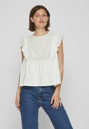 Top de algodón blanco con detalles de encaje y mangas con volantes; corte suelto y cintura fruncida, combinado con jeans de mezclilla azules.
