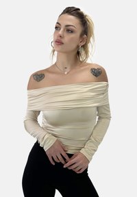 Cremefarbenes Oberteil mit schulterfreiem Design und langen Ärmeln, das eine Raffung über der Brust aufweist. Das Model posiert in schwarzen, taillierten Hosen.