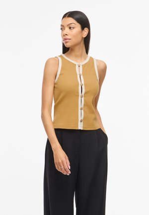 Femme aux longs cheveux foncés portant un haut sans manches moutarde avec bordure beige et boutons décoratifs, associé à un pantalon large noir.