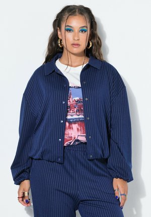Frau mit langen Haaren, die zu Zöpfen gestylt sind, trägt eine blaue Nadelstreifenjacke und Hose, ein grafisches T-Shirt, große Creolen und kräftigen blauen Lidschatten.