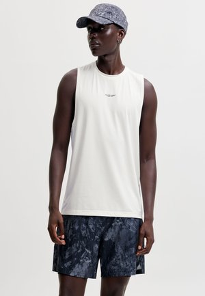 Personne à la peau foncée portant un débardeur blanc, un short sombre à motifs et une casquette de baseball grise à motifs, debout devant un fond uni.