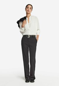 Vrouw in witte blouse die in zwarte op maat gemaakte broek is gestopt, met een zwarte riem, terwijl ze een zwarte jas over haar schouder houdt, staand tegen een witte achtergrond.