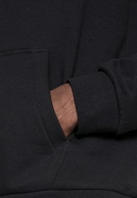 Sudadera negra con capucha, que presenta un bolsillo frontal y puños de canalé. La tela parece suave con una textura lisa. La mano está reposando en el bolsillo.