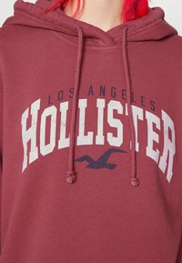 Sudadera color burdeos con un bolsillo canguro en la parte frontal y cordones ajustables. Presenta un texto gráfico en blanco y azul marino que dice "HOLLISTER LOS ÁNGELES."