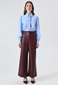 Camicia blu con maniche a sbuffo e dettaglio a fiocco abbinata a pantaloni in pelle bordeaux a gamba larga e décolleté bordeaux a punta.