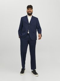 Marineblauer Anzug mit taillierter Passform, einreihiger Blazer mit zwei Knöpfen und Hosen mit flachem Vorderteil. Getragen mit einem weißen Hemd und schwarzen Sneakers.