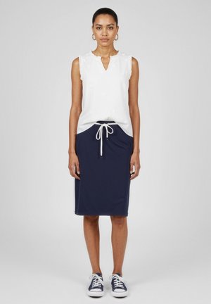 Donna con una camicetta bianca senza maniche, gonna blu navy a ginocchio con coulisse e sneakers blu navy, in piedi davanti a uno sfondo bianco semplice.