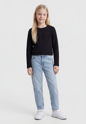 Blondínka v čiernom tričku s dlhým rukávom, svetlomodrých džínsach a bielych teniskách stojí pred jednoduchým svetlým pozadím a usmieva sa.