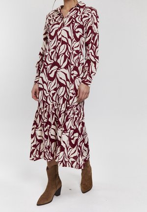 Kobieta ubrana w sukienkę midi z długim rękawem, ozdobioną bordowo-białymi, abstrakcyjnymi wzorami liści, w połączeniu z brązowymi zamszowymi butami sięgającymi połowy łydki.