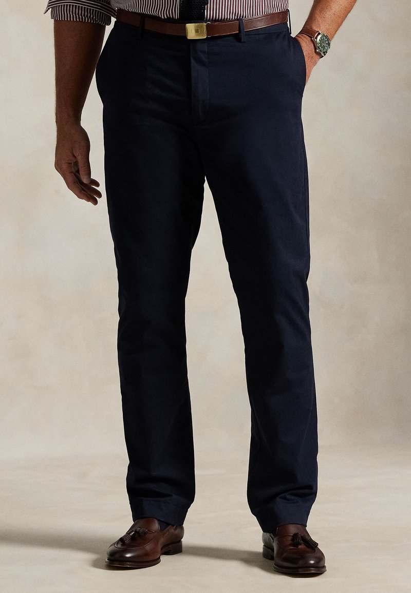 Férfi sötétkék nadrágban, barna bőr övvel, csíkos ingben, karórával és barna bojtos mokaszinban, kezét a zsebében tartva áll.