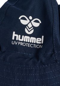 Marinblå bucket hat med en slät textur, med en framträdande vit logotyp och texten "UV SKYDD". Vadderad detaljering längs skärmen.