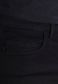 Zwarte spijkerbroek met een klein voorvakje, zichtbare stiksels en een gladde, licht gestructureerde stof.