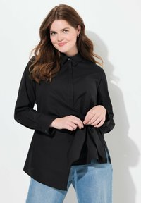 Camisa negra de botones, con la cintura atada, mangas largas y cuello clásico; textura suave, combinada con jeans de color azul claro.