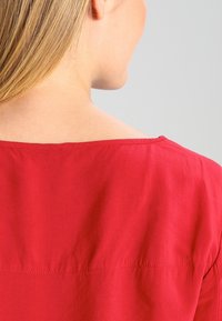 Chemise rouge avec une texture lisse, présentant un décolleté large et une couture subtile le long du dos. Le tissu semble léger et respirant.