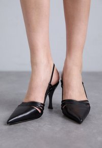 Svarta läderklackar med slingback-design och spetsig tå, med korsande remmar över foten och en tunn, böjd klack. Slät yta.
