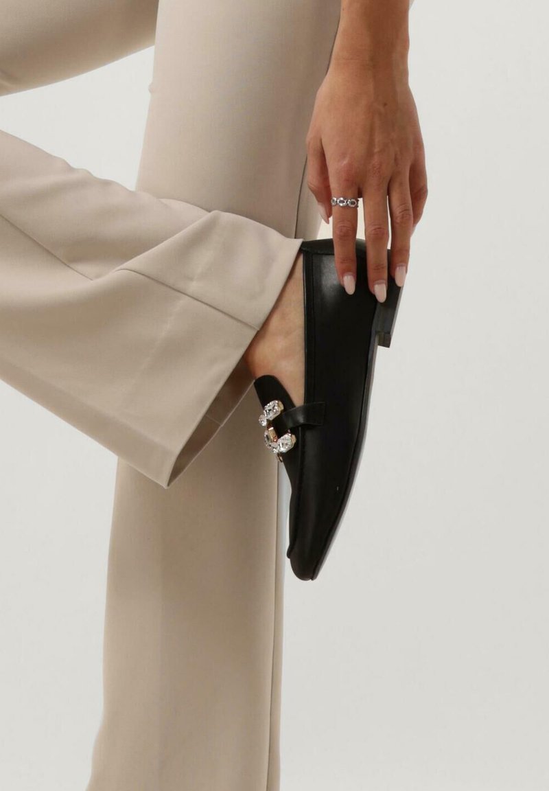 Zwarte leren instappers met een lage hak, een decoratieve band met metalen accenten en een puntige neus. Ggedragen met beige broeken.