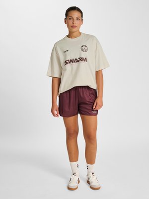Jonge vrouw staand, draagt een beige oversized overhemd met logo’s, donkerrode sportshorts, witte sokken en witte sneakers op een effen achtergrond.