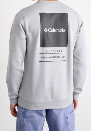 Mann trägt hellgrauen Columbia-Sweatshirt mit schwarzem quadratischem Logo und dem Text „Since 1938 Portland, Oregon, USA“ auf dem Rücken.