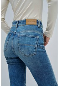 Mujer vistiendo unos jeans de mezclilla Salsa azules vistos desde atrás, combinados con una blusa de manga larga beige, con las manos apoyadas en las caderas.