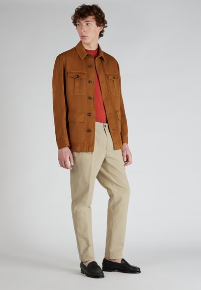 Brązowa kurtka zapinana na guziki z czterema kieszeniami z przodu, czerwona koszula pod spodem, beżowe spodnie i czarne mokasyny, na modelu na neutralnym tle.