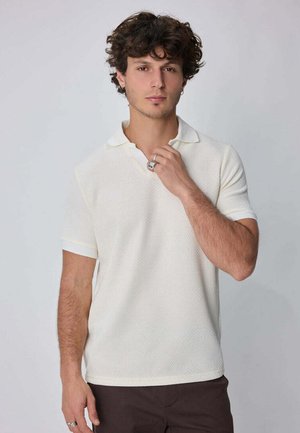 Jeune homme aux cheveux bouclés portant un polo blanc à manches courtes et un pantalon sombre, tenant son col d'une main.