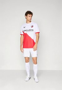 Camisa de futebol branca e vermelha de mangas curtas, com um padrão diagonal vermelho e marcação preta. Combina com calções e meias brancos.