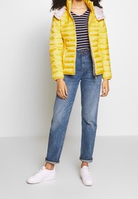 Giacca a vento gialla con colletto grigio chiaro, abbinata a una maglietta a righe bianche e nere, jeans azzurri chiari e sneakers bianche.