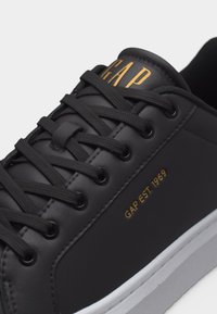 Μαύρο δερμάτινο αθλητικό παπούτσι με μαύρα κορδόνια, χρυσό λογότυπο "GAP" στη γλώσσα και "GAP EST. 1969" σε χρυσό στη πλευρά πάνω από τη λευκή σόλα.