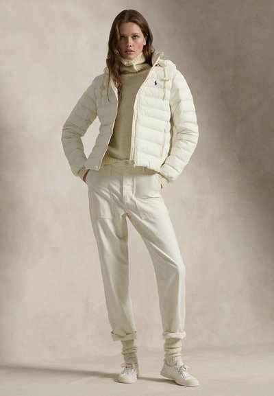 Vrouw staat met handen in de zakken, draagt een crèmekleurige donzen jas, een beige coltrui, losse broek, sokken en witte sneakers.