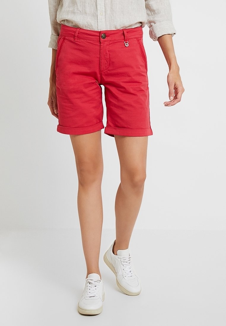 Pantalones cortos de algodón rojos con puños doblados, que cuentan con bolsillos frontales y un cierre de botón. Combinados con zapatillas blancas y una camisa de color claro.