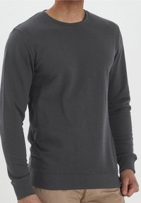 Hombre que lleva un sudadera de cuello redondo de manga larga gris oscuro y pantalones beige, mostrado de cuello a cintura sobre un fondo liso.