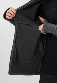 Personne portant une chemise noire tient ouverte une veste à fermeture éclair côtelée foncée, montrant une grande poche intérieure et un trou pour le pouce au niveau du poignet.