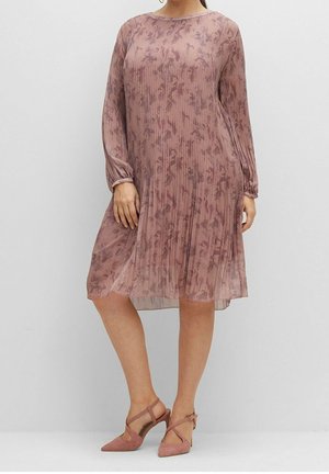 Vrouw draagt een knielange jurk met lange mouwen in stoffig rozenroze, geplooid met een abstract bloemmotief, en bijpassende pumps met spitse neus en bandjes.