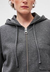 Šedá zapínacia mikina vyrobená z mäkkej tkaniny, s prednou vreckom, sťahovacími šnúrkami a strieborným zipsom s špicatou ťahacou slučkou.