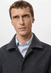 Cappotto in lana grigio scuro con una camicia a righe blu e bianche. Il cappotto presenta un colletto con risvolto e chiusura con bottoni, con una finitura testurizzata.