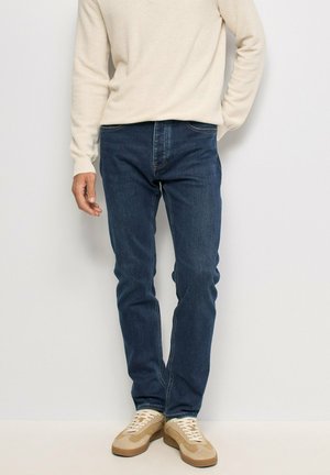 Hombre con suéter crema, vaqueros azul oscuro y zapatillas casuales beige de pie contra una pared blanca.