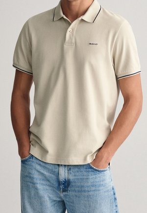 Beżowa koszulka polo o dopasowanym kroju, z krótkimi rękawami i czarnym obszyciem wokół kołnierzyka i rękawów. Na piersi znajduje się logo GANT.