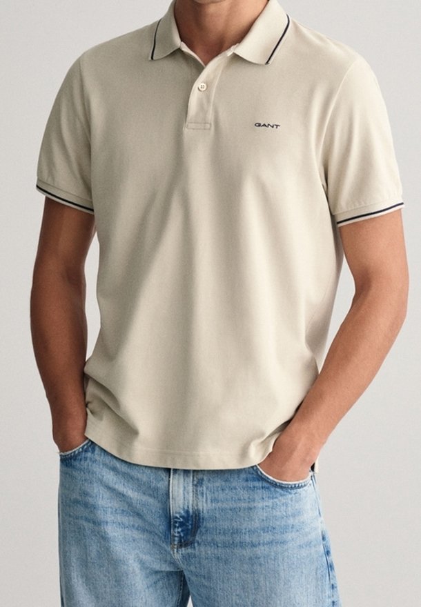 Polo beige à coupe slim, manches courtes, avec un liseré noir autour du col et des manches. Logo GANT sur la poitrine.