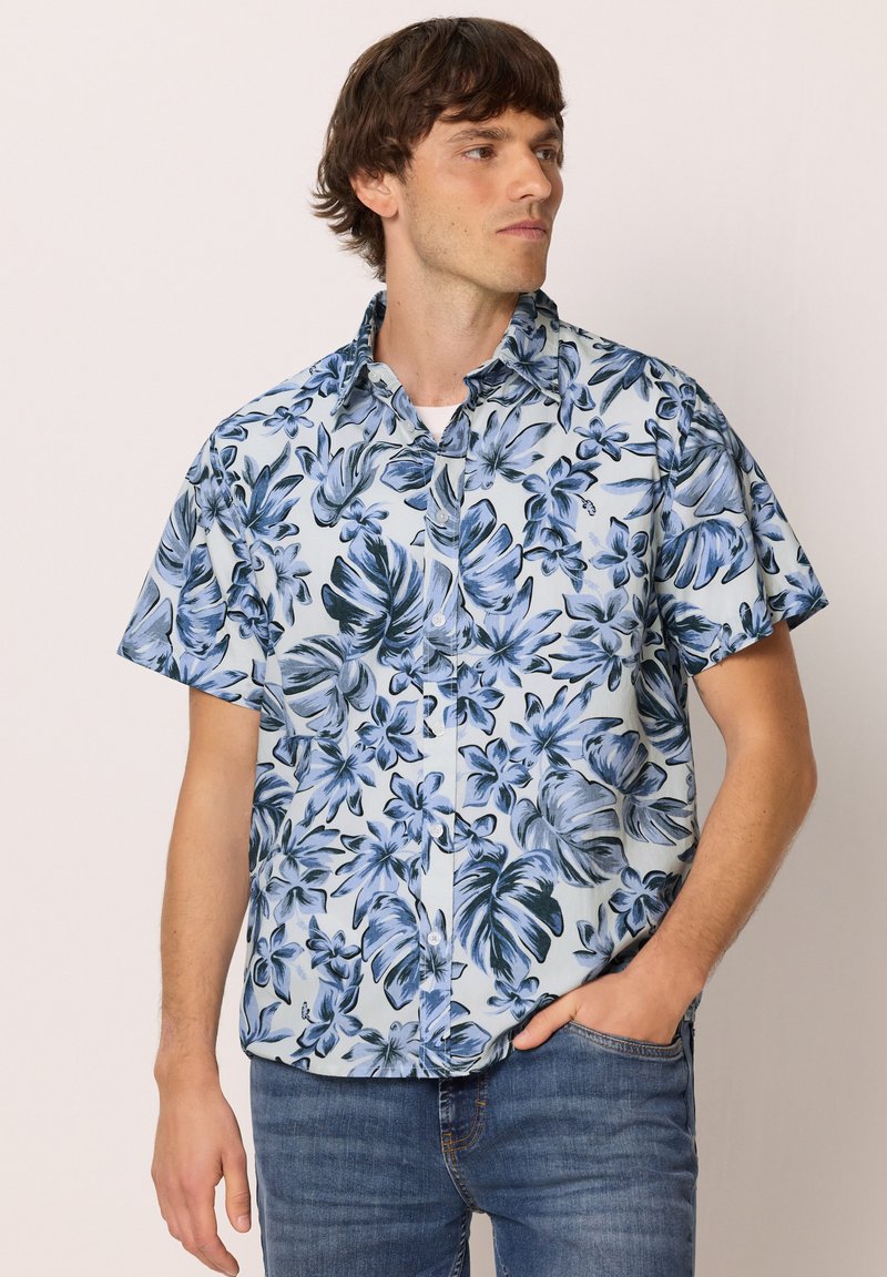 Homme portant une chemise boutonnée bleu et blanc à manches courtes avec un grand imprimé floral, debout, une main dans la poche du jean, regardant vers la droite.