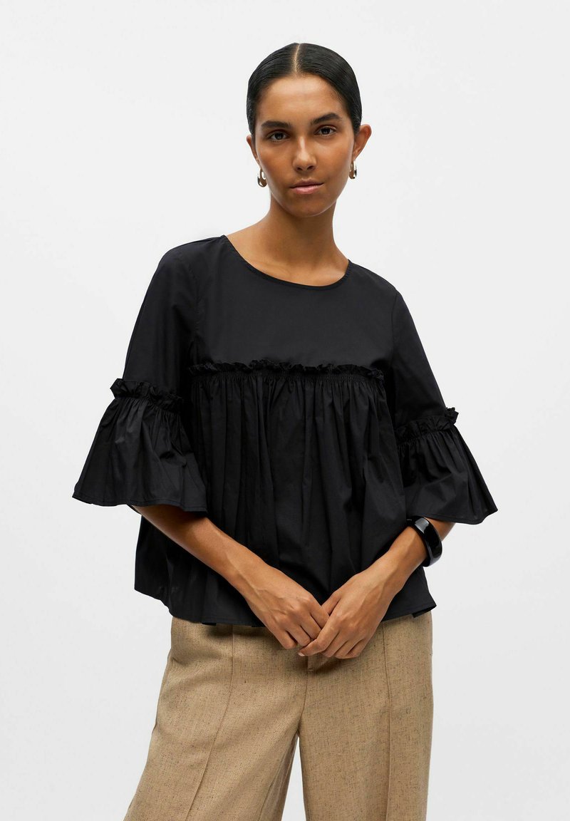 Must monochrome top, millede lõhikutega varrukad ja kogunenud esiosa. Sile kangas ümmarguse kaelusega, mis on paaritatud beežide laia säärega pükstega.
